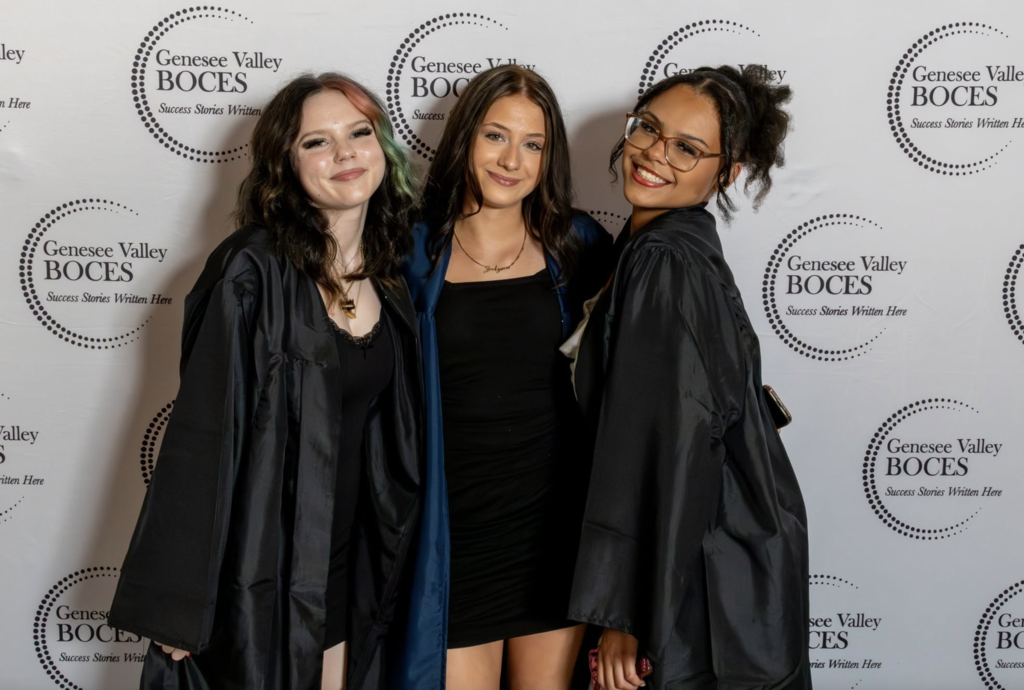 Three students stand in front of a step and repeat banner with gowns on.  
