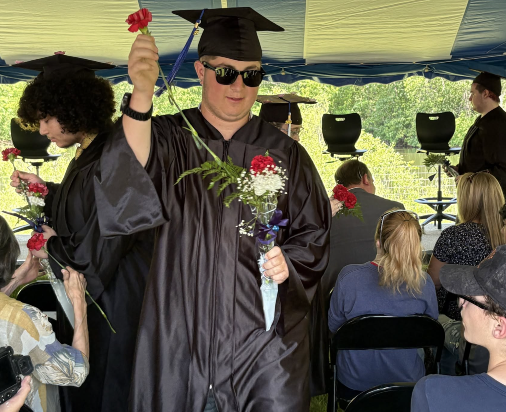 A student with a hand full of flowers getting ready to hand one out.  He is wearing a cap, gown, and sunglasses. 