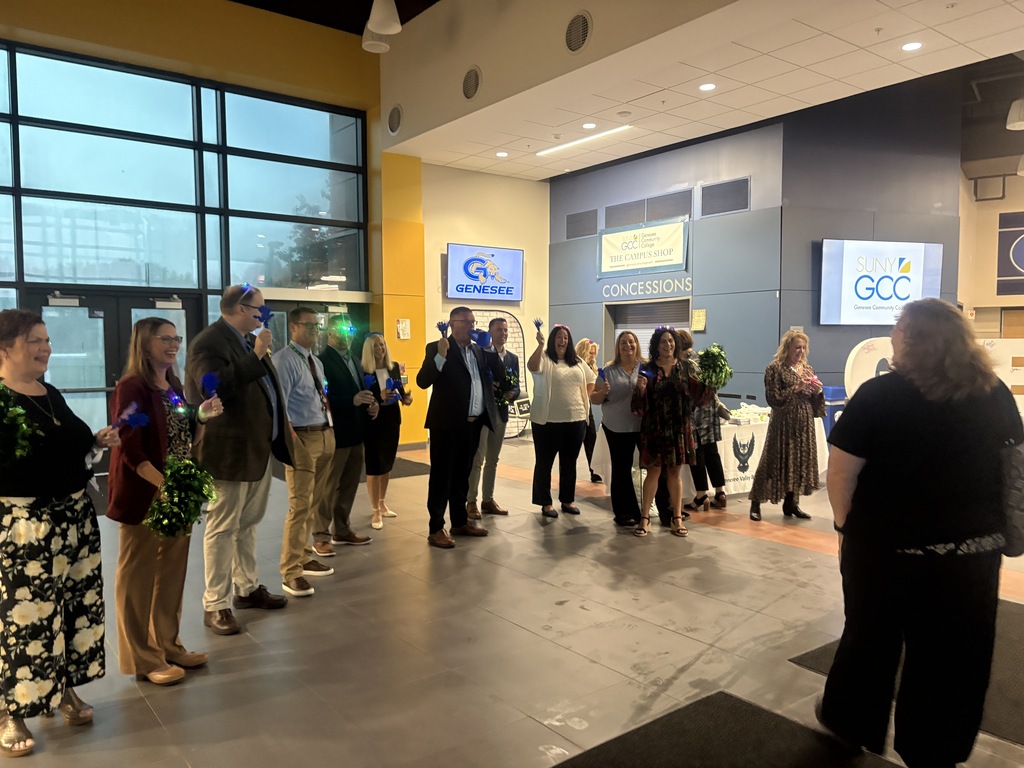 A group of people stand in a line inside a modern building with large windows. Monitors with logos hang on the wall.