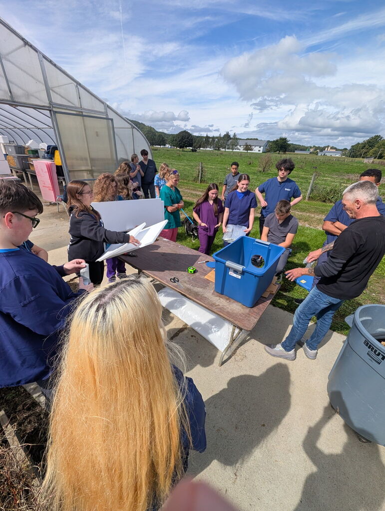 Group of people in a greenhouse. A table has a blue container with an item inside.