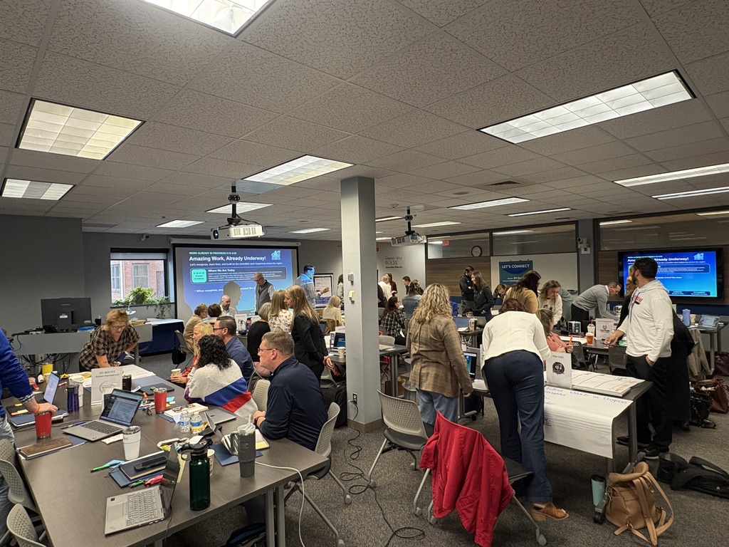 People gathered in a room with a projector screen, tables, chairs, and bags. Laptops and items are on tables.