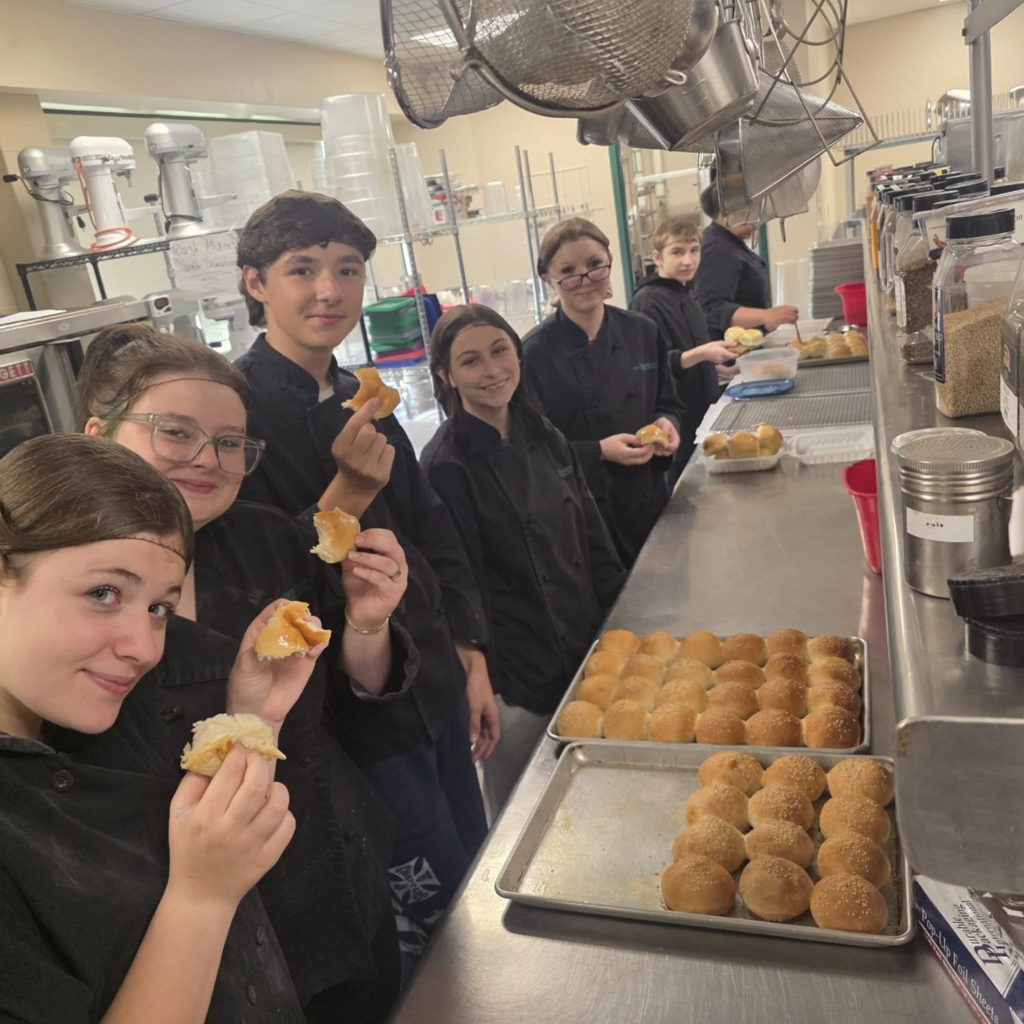 A group of people, some holding food, stand in a kitchen. Trays of bread and a metal countertop are visible.