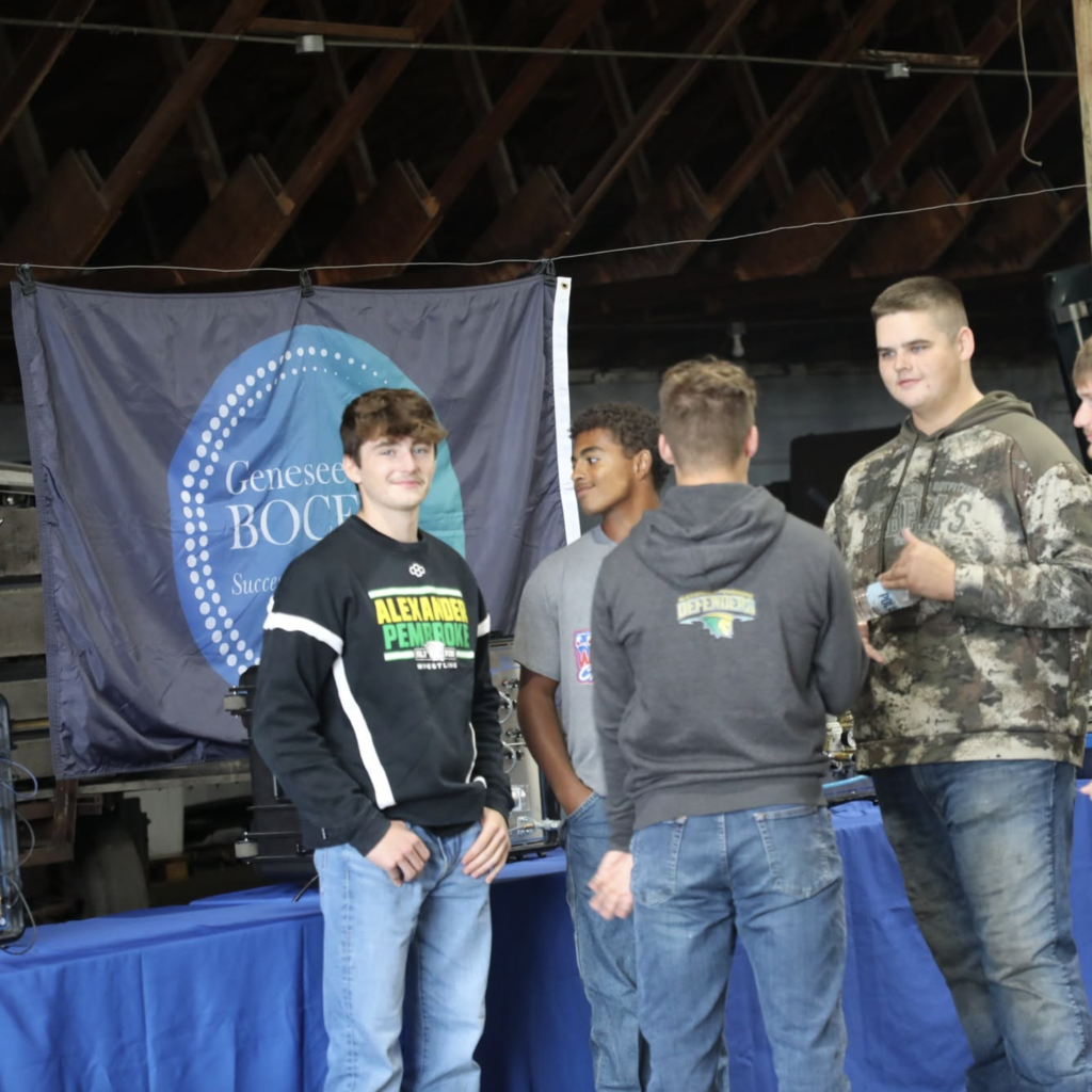 Five boys in jeans and hoodies stand in front of a table and a banner.