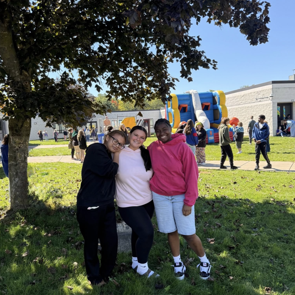 Three people standing close together, smiling, on a green lawn under a tree with an inflatable structure in the background.