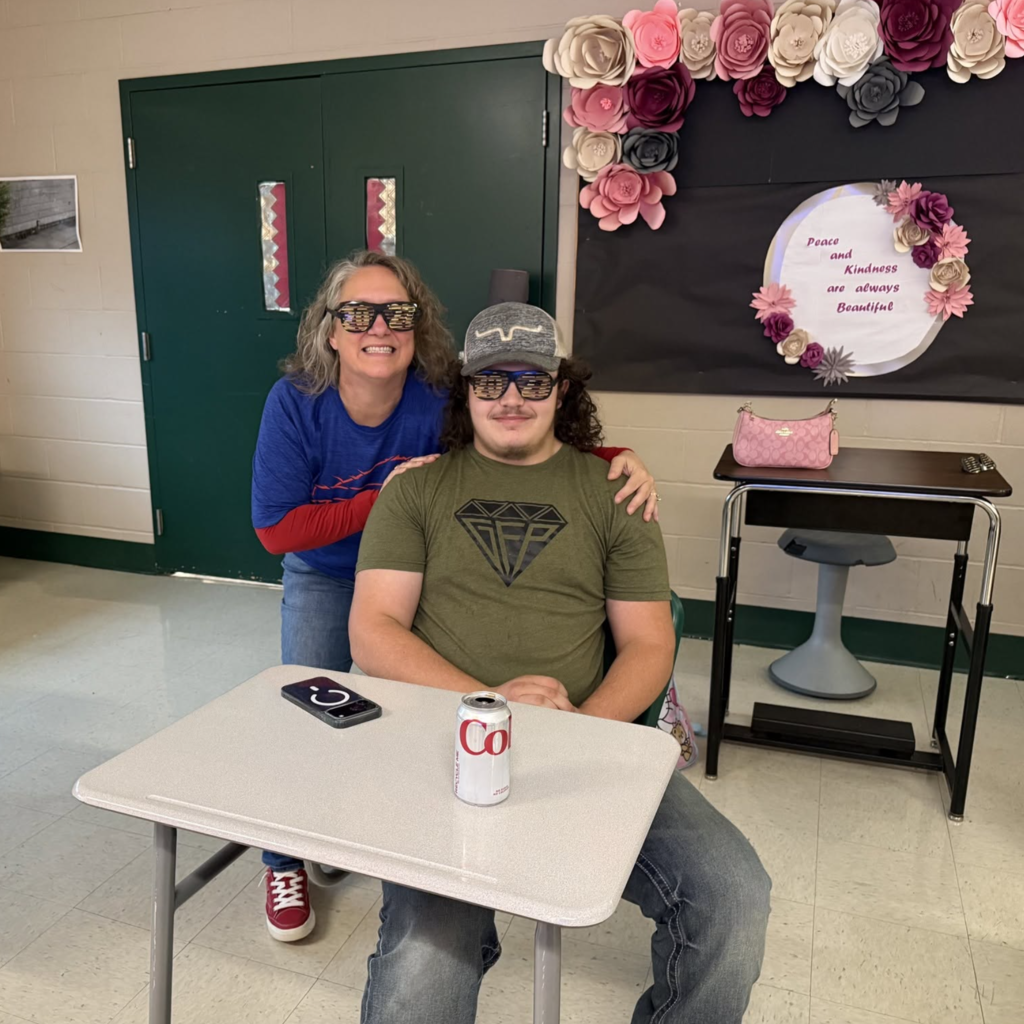 Two people seated near a desk with a can of soda; one is wearing glasses, the other a cap.