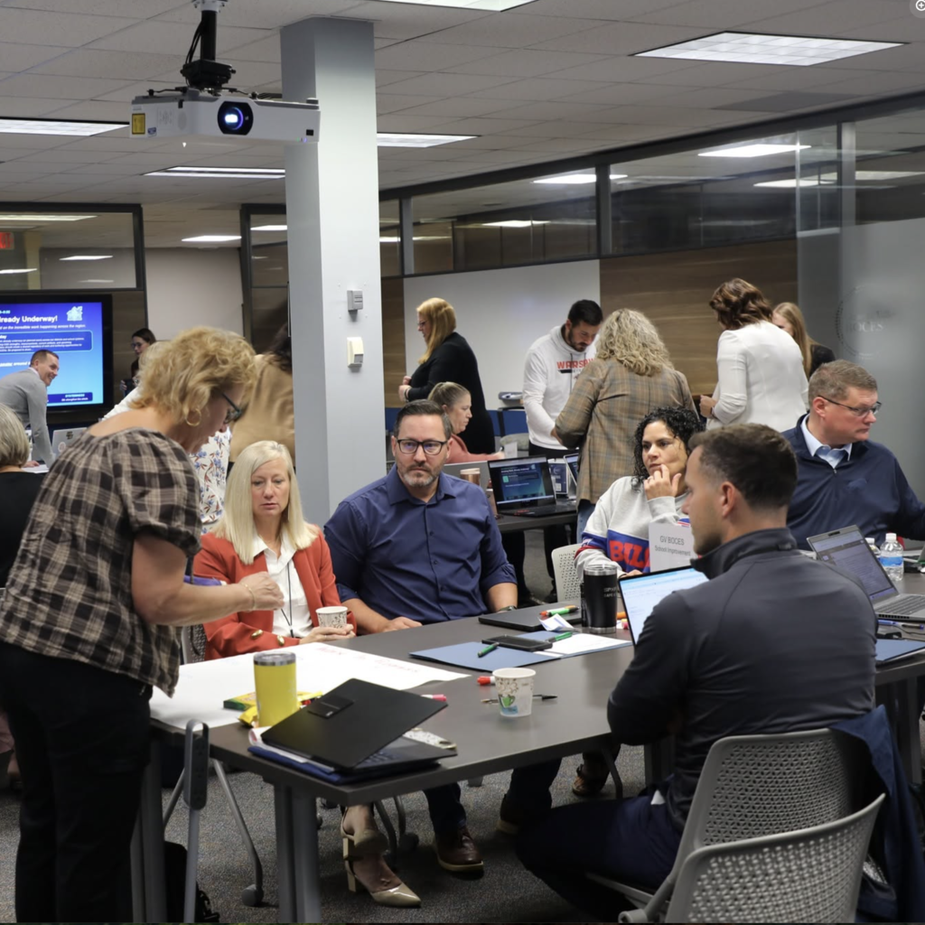People in a room with tables. Some are seated, others standing. Laptops, cups, and papers on tables.