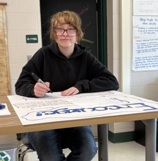 A student sits at a desk writing on a large poster with a marker.