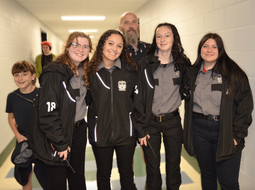 A group of students wearing jackets stand in a hallway with white walls, one holding a wand.