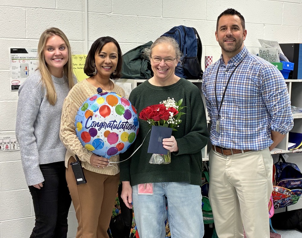 Four staff members stand together indoors, smiling for a photo. The person second from the right holds a bouquet of red roses and a card, while another holds a colorful “Congratulations” balloon. Coats, cubbies, and school supplies are visible in the background, suggesting a school setting.