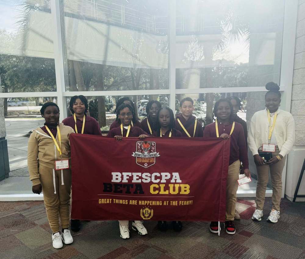 A group of Brown’s Ferry Elementary School students stand indoors holding a maroon banner that reads “BFESCPA Beta Club – Great Things Are Happening at the Ferry!” The students are wearing school uniforms and name badges, smiling proudly while posing together in front of large windows with trees visible outside.