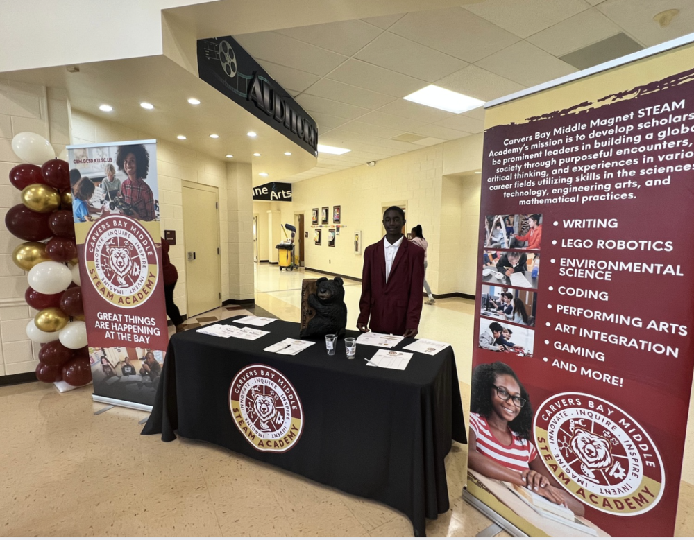 Hello Bear Family! This is a picture of one of our Bear Ambassadors standing behind a table in front of the auditorium. The school's Magnet banners are standing g on both sides of the table. There are gold, burgundy, and white balloons adorning one of the banners. There are sign-in sheets and programs on the table also.