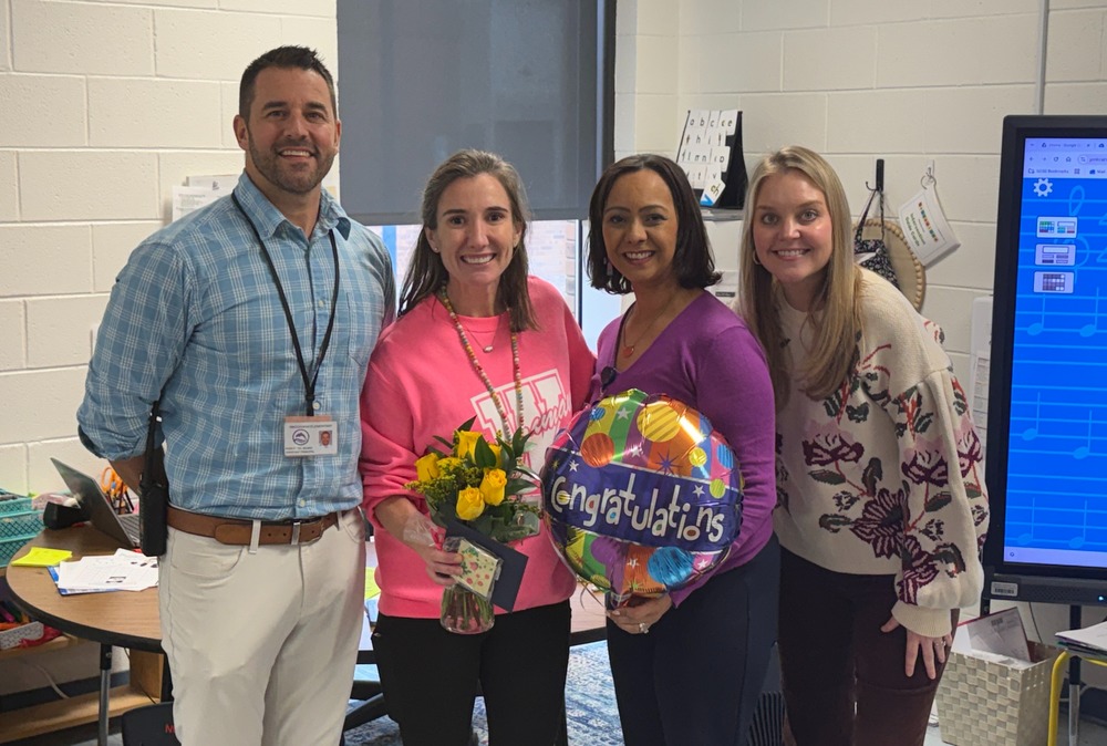 Four staff members stand together in a classroom smiling for a photo. The person second from the left holds a bouquet of yellow flowers while another holds a colorful “Congratulations” balloon. Classroom furniture, student materials, and a large screen display are visible in the background.