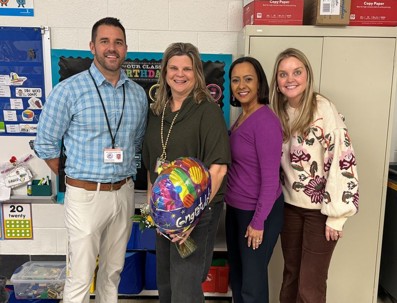 Four staff members stand together in a classroom smiling for a photo. The person in the center holds a colorful congratulatory balloon and flowers while the others stand beside her in celebration. Classroom materials, posters, and storage cabinets can be seen in the background.