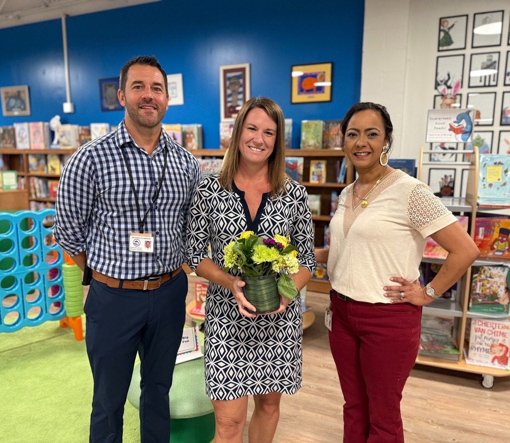 Three smiling adults in a school library, with the center person holding a small bouquet of flowers.