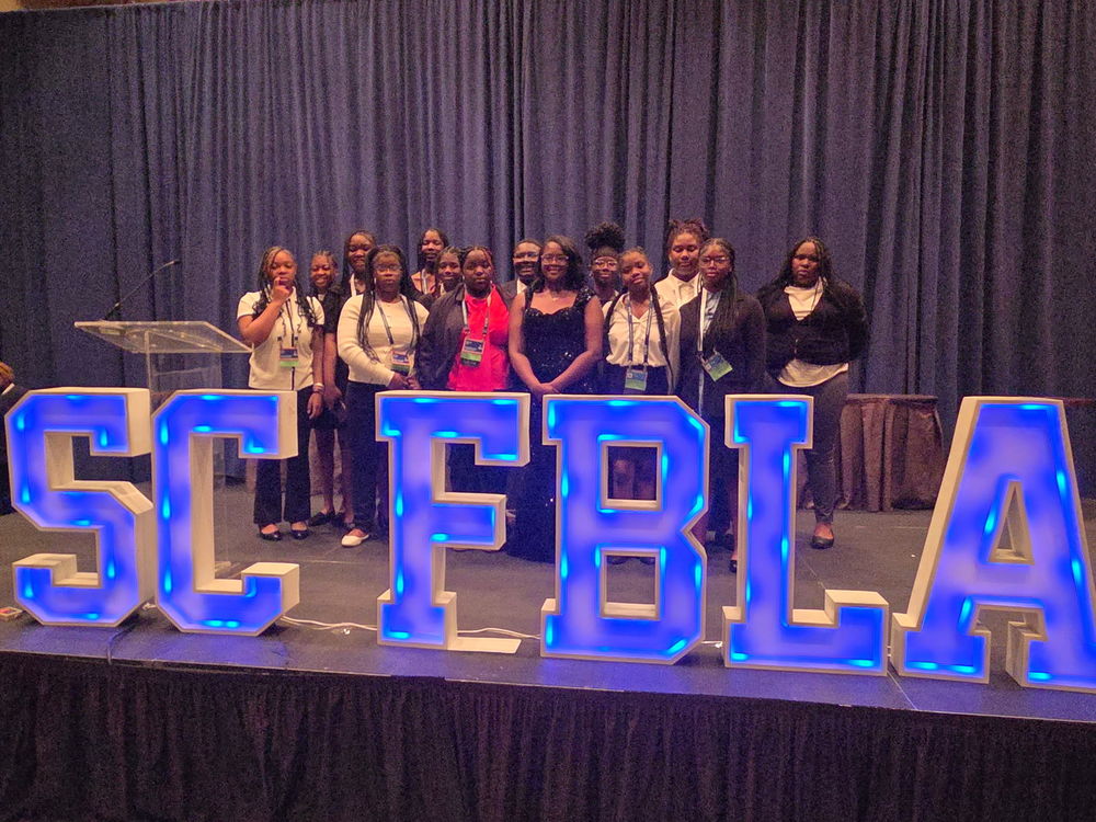 CBMSA FBLA students standing on stage behind d a blue lit-up SC FBLA sign.