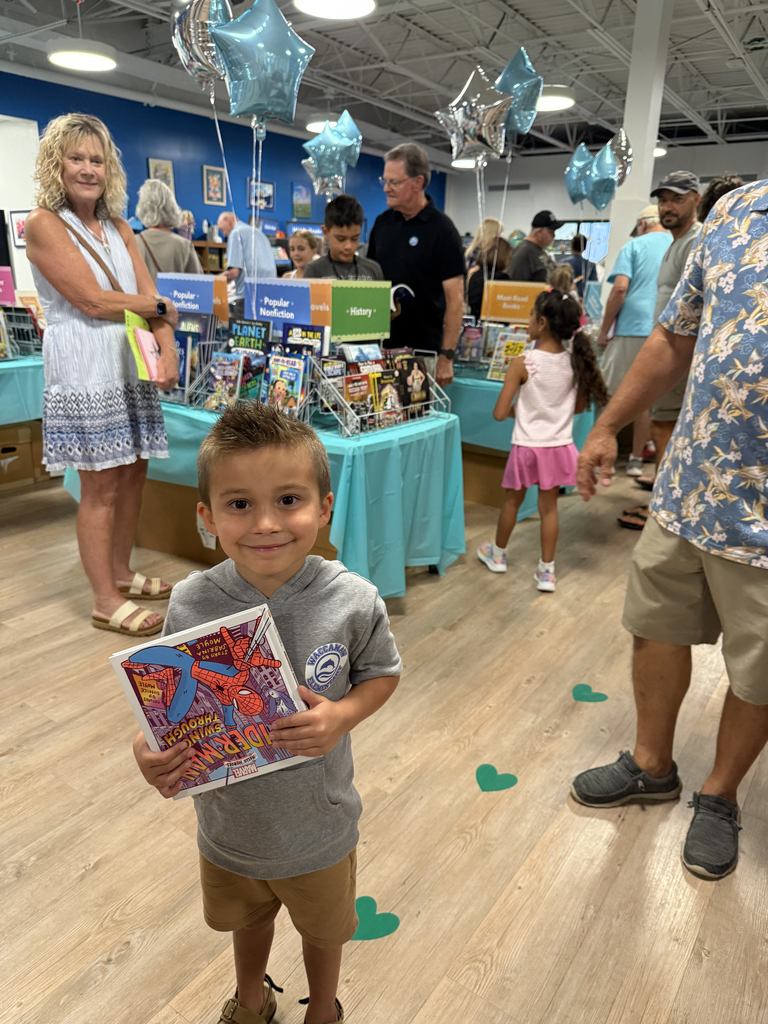 A young boy holding a book with tables covered in books behind him