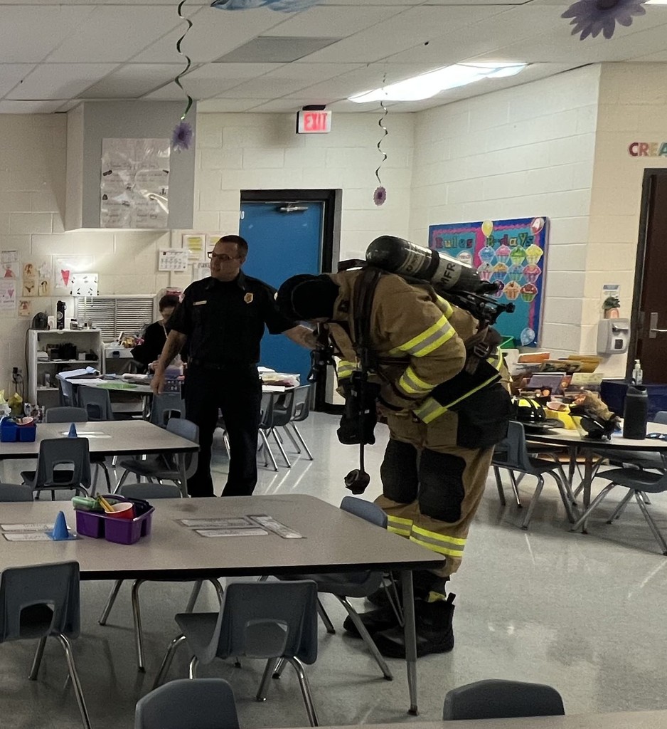 Two firefighters stand in a classroom; one is speaking while the other demonstrates putting on protective gear.