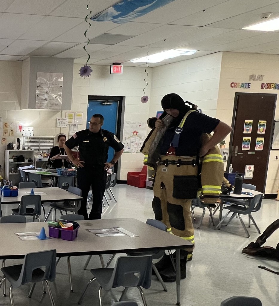 Two firefighters stand in a classroom; one is speaking while the other demonstrates putting on protective gear.