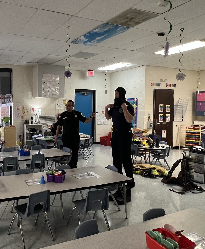 Two firefighters stand in a classroom; one is speaking while the other demonstrates putting on protective gear.