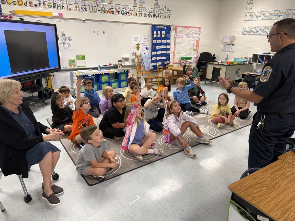 A firefighter speaks to a group of young students sitting cross-legged on the classroom floor, with several children raising their hands to answer questions.