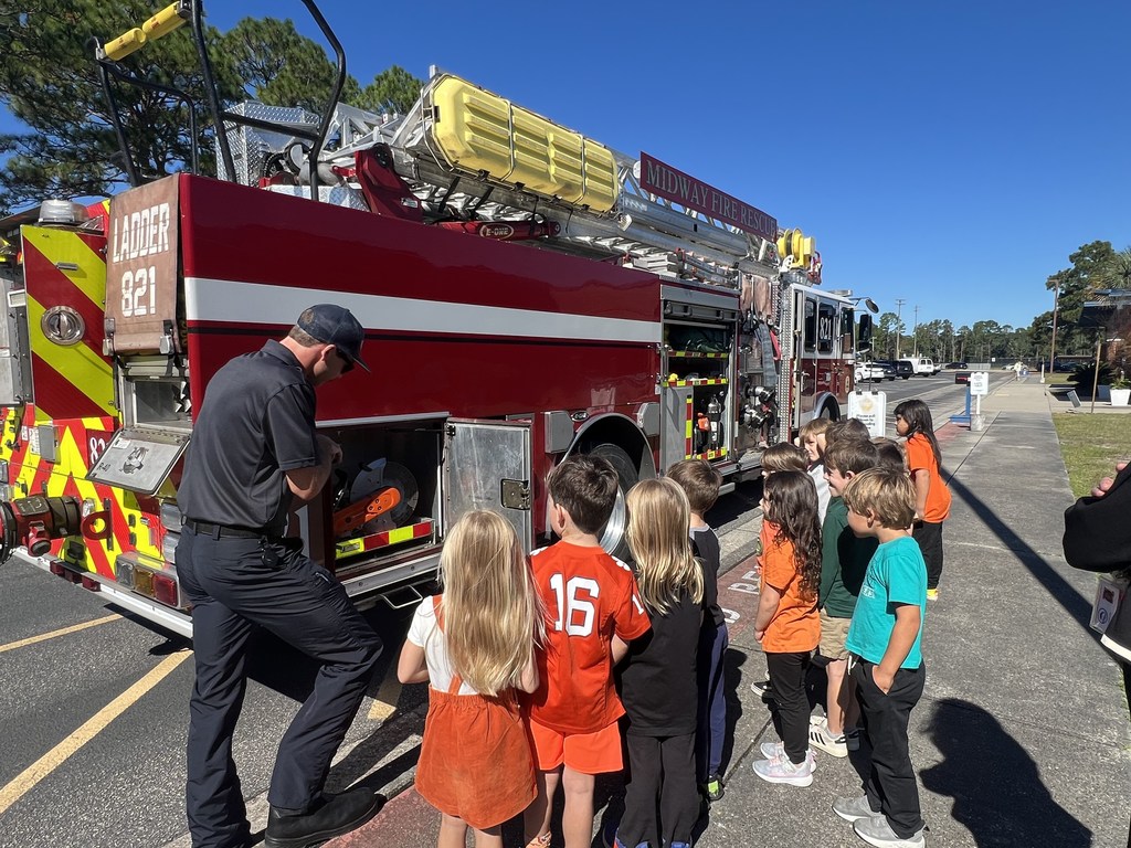A firefighter shows equipment from a fire truck to a group of elementary students standing outside in the sunlight. The truck is labeled “Midway Fire Rescue,” and the children watch closely, lined up along the side of the vehicle.