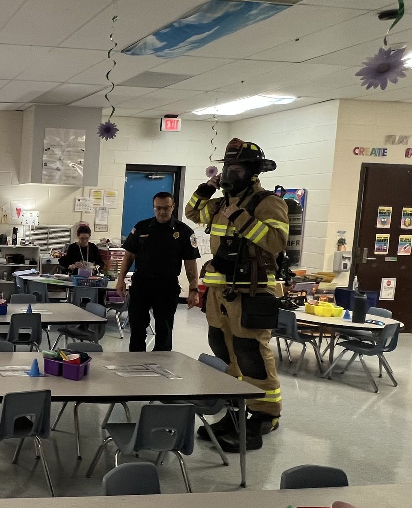 Two firefighters stand in a classroom; one is speaking while the other demonstrates putting on protective gear.