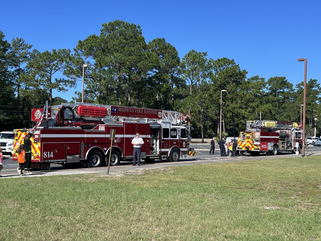 Two Midway Fire Rescue trucks are parked outside on a sunny day as firefighters and students gather around them.
