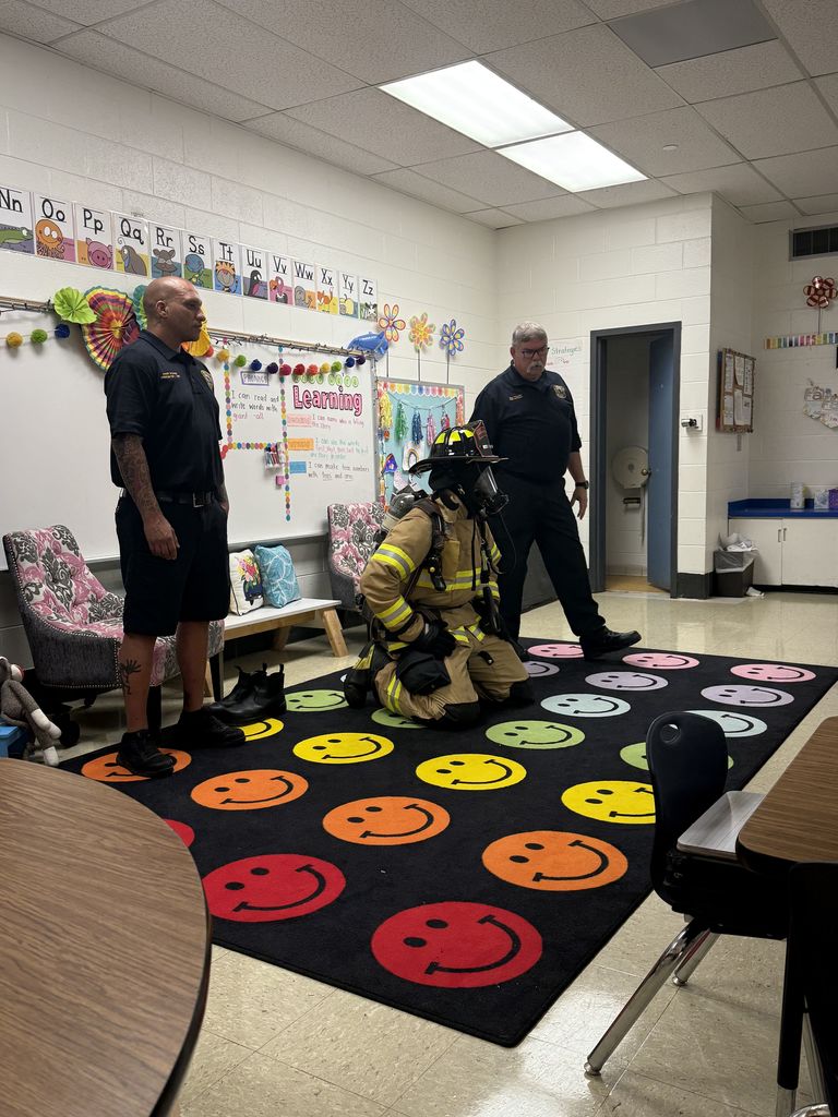 Firefighters give a classroom presentation as one kneels on a colorful rug wearing full firefighting gear while two others stand nearby explaining equipment.