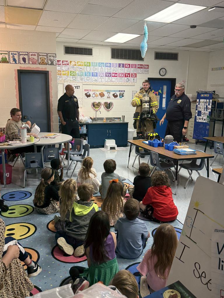 Firefighters speak to a group of young students seated on a colorful classroom rug as one firefighter demonstrates wearing full turnout gear.