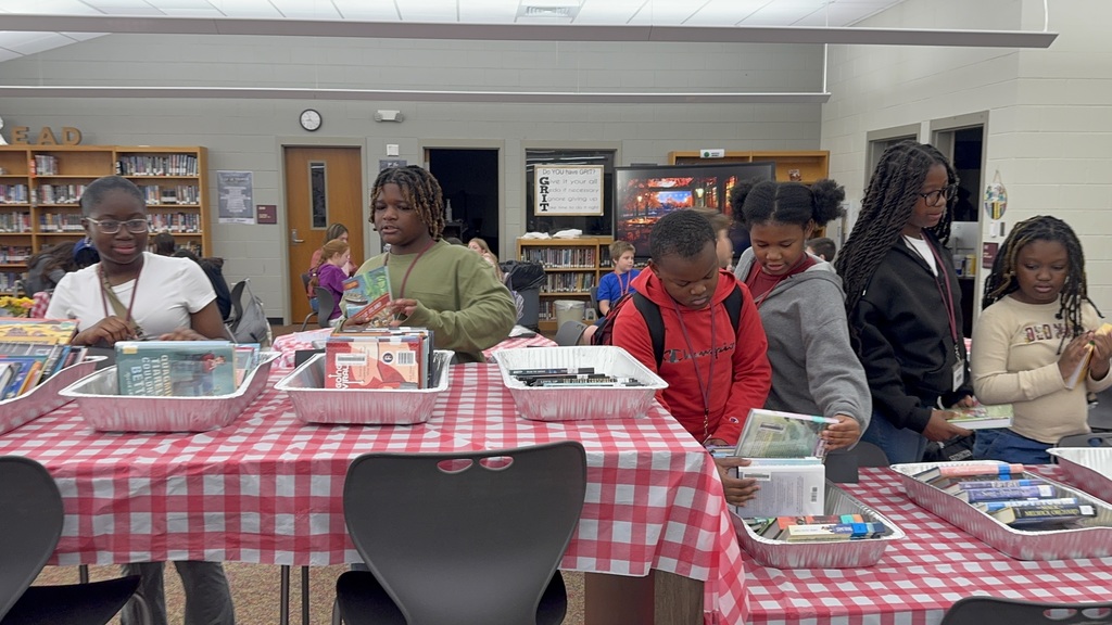 Kids are standing at a table with a red-checkered tablecloth , looking at books.
