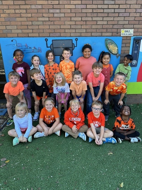 Boys & Girls sitting on a green carpet together, most wearing the color orange. 