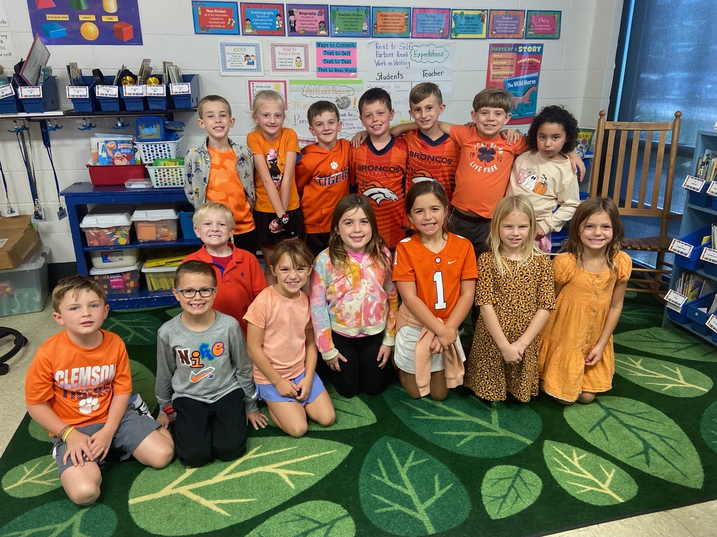 Boys & Girls sitting on a green carpet together, most wearing the color orange. 