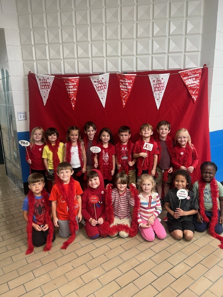 A group of young students dressed in red pose in front of a red backdrop decorated with “Be Drug Free” and “Celebrate Red Ribbon Week” banners. The children smile and hold props supporting Red Ribbon Week’s drug-free message.