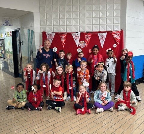 A group of elementary school students and their teacher posing together in a hallway in front of a red backdrop decorated with banners that say “Team Up Against Bullies and Drugs.” The children are smiling and holding red props, such as pom-poms and signs, as part of a themed school spirit or awareness event.