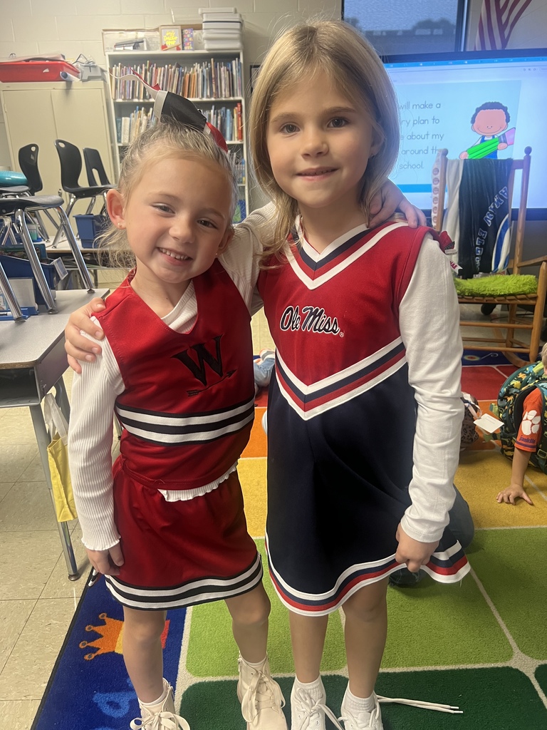 Two young girls in cheerleading uniforms smiling and standing arm in arm in a classroom.