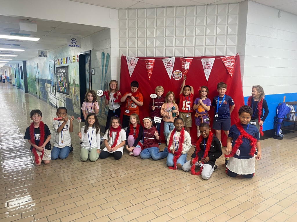 A group of elementary school students posing together in a hallway in front of a red backdrop decorated with banners that say “Team Up Against Bullies and Drugs.” The children are smiling and holding red props, such as pom-poms and signs, as part of a themed school spirit or awareness event.