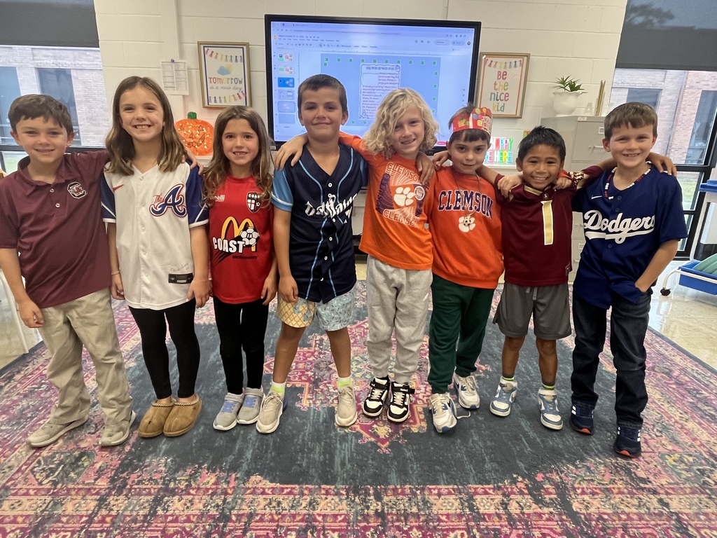A group of eight smiling elementary school children standing side by side in a classroom, each wearing a sports or team shirt, with their arms around each other’s shoulders.