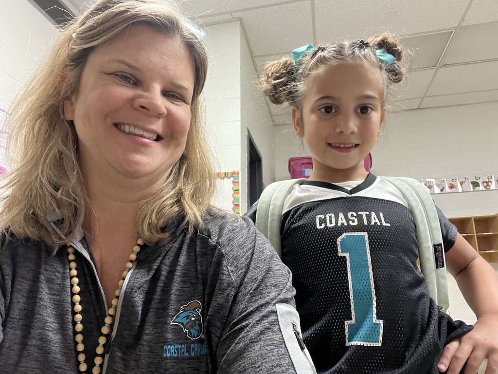 A smiling teacher and young student posing together in a classroom. Both are wearing black and teal Coastal Carolina Chanticleers shirts.