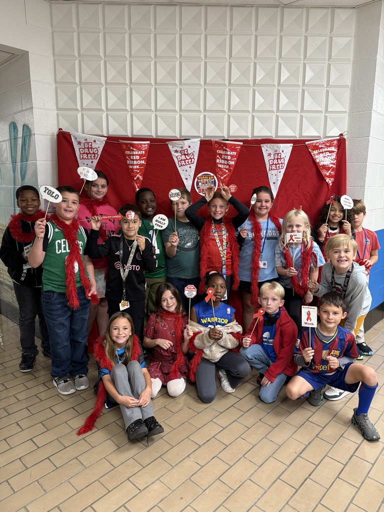 A group of elementary school students posing together in a hallway in front of a red backdrop decorated with banners that say “Team Up Against Bullies and Drugs.” The children are smiling and holding red props, such as pom-poms and signs, as part of a themed school spirit or awareness event.
