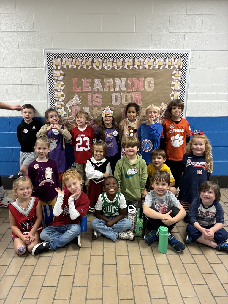 A group of kindergarten students smiling and posing and wearing sports-themed shirts and jerseys representing various teams and colors, standing and sitting together in two rows in a school hallway.