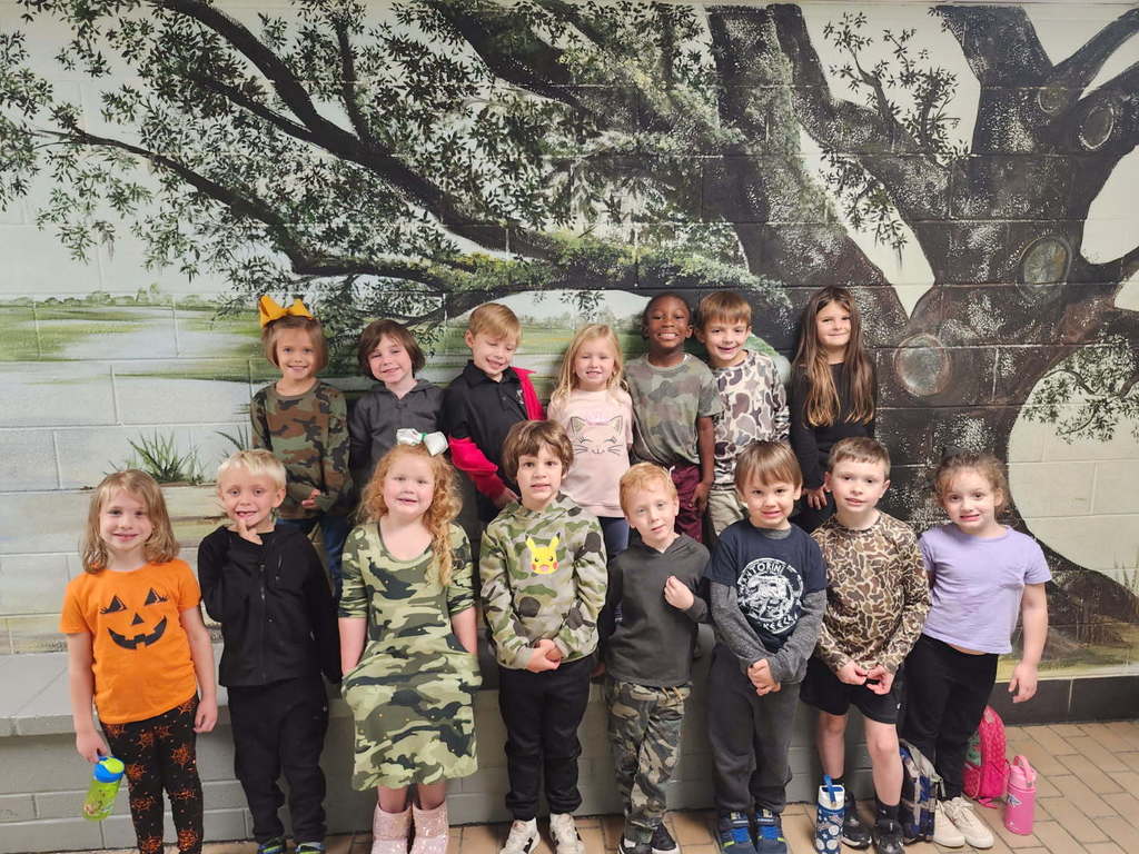 A group of children stand indoors in front of a mural of a large tree, smiling and wearing a mix of camouflage and colorful outfits.