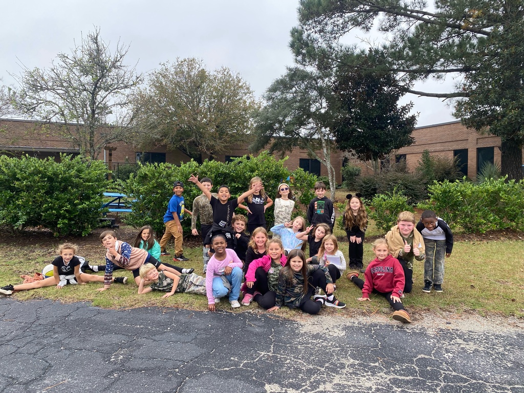 A large group of children pose and smile outside near bushes and trees in front of a brick school building.