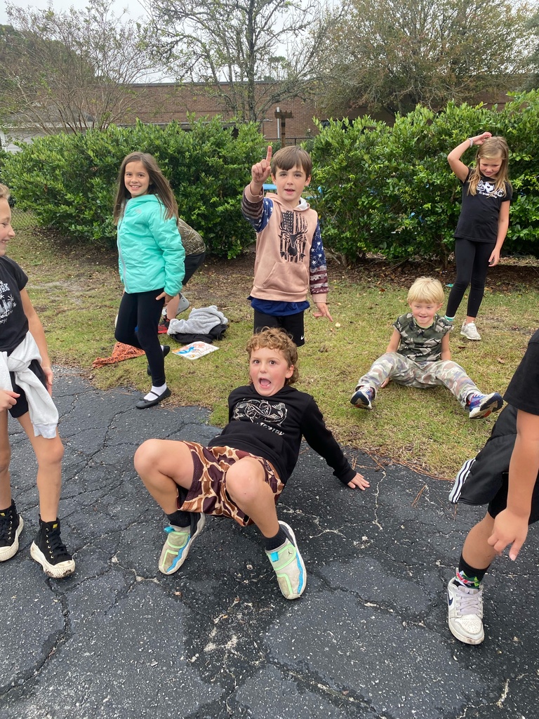 A group of children play outside near bushes, with one boy crab-walking on the pavement and others standing or sitting around him.