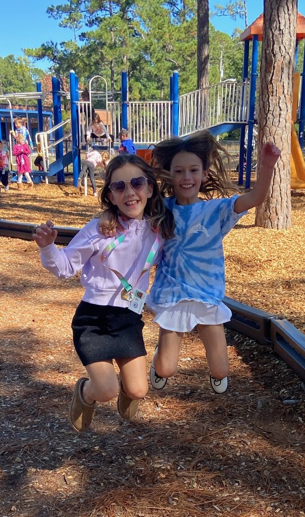 Two girls jump joyfully in the air on the playground, smiling as their hair flies up.
