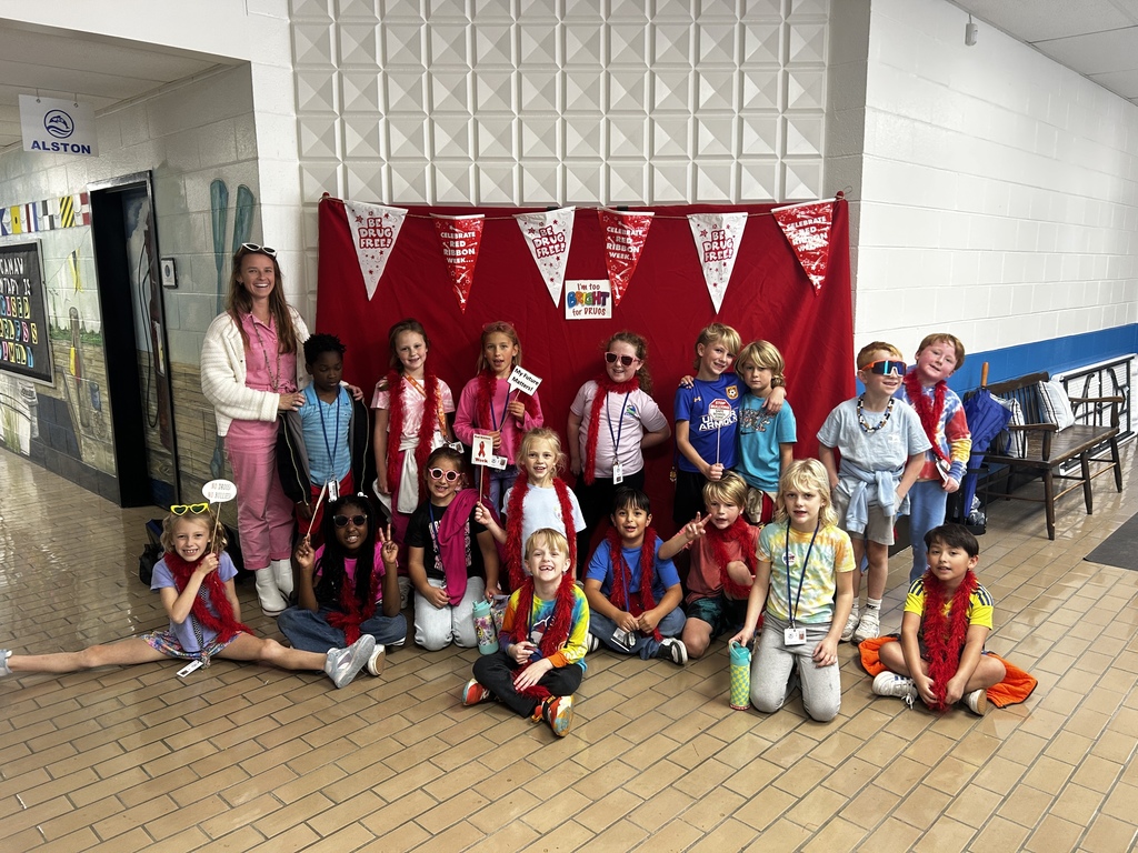 A teacher and her class pose in front of a red photo backdrop decorated with Red Ribbon Week banners that say “Be Drug Free.” The teacher and students wear colorful outfits and sunglasses, and several children hold props with anti-drug messages. Everyone is smiling and having fun in a bright school hallway.