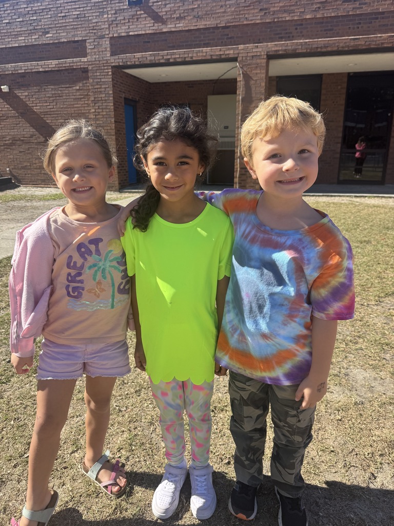 Three young students smile and pose together outdoors in front of a brick school building. They are dressed in colorful clothing—one wearing pink, another in a bright neon green shirt, and the third in a tie-dye shirt.