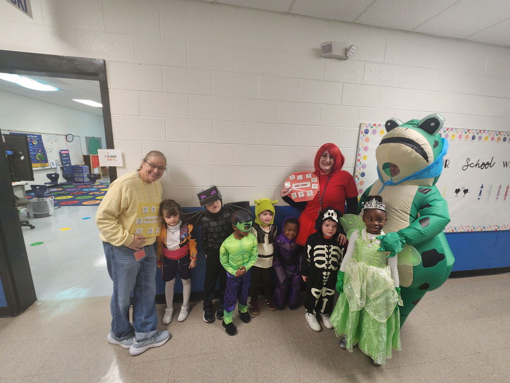 Two teachers and a group of young students stand in a hallway wearing Halloween costumes. Outfits include a skeleton, princess, frog, superhero, and red-haired character. The teachers are dressed as emotion-themed characters with labeled signs, smiling together against a white and blue wall.