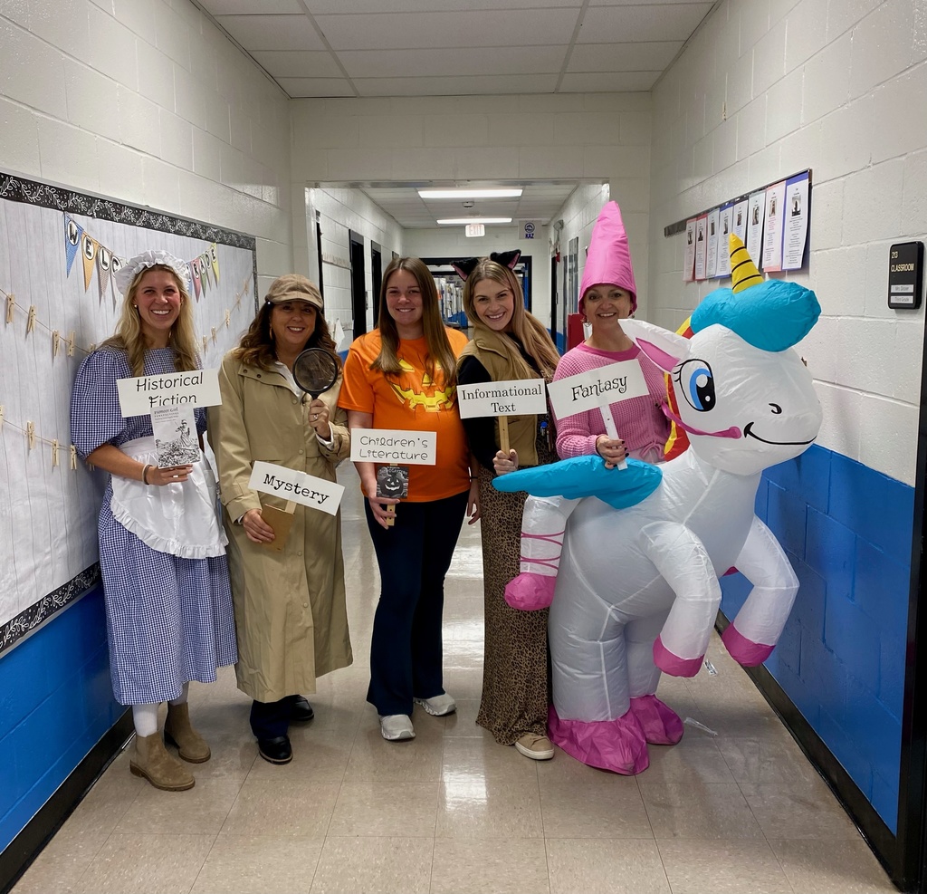 Five teachers pose in a school hallway dressed to represent different book genres. They hold signs labeled “Historical Fiction,” “Mystery,” “Children’s Literature,” “Informational Text,” and “Fantasy.” One teacher wears an inflatable unicorn costume, and another is dressed as a detective holding a magnifying glass.