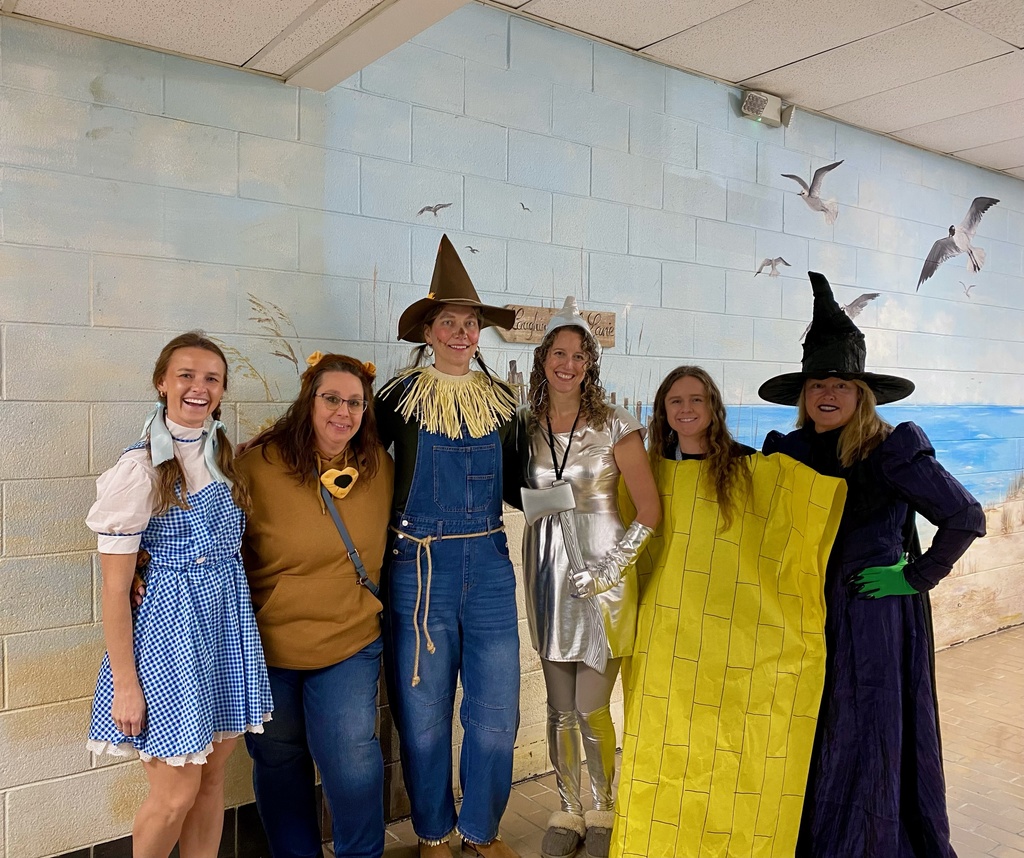 Six teachers dressed as characters from The Wizard of Oz stand smiling in a school hallway. Costumes include Dorothy in a blue gingham dress, the Cowardly Lion, the Scarecrow, the Tin Man, the Yellow Brick Road, and the Wicked Witch of the West. A beach mural with seagulls decorates the wall behind them.