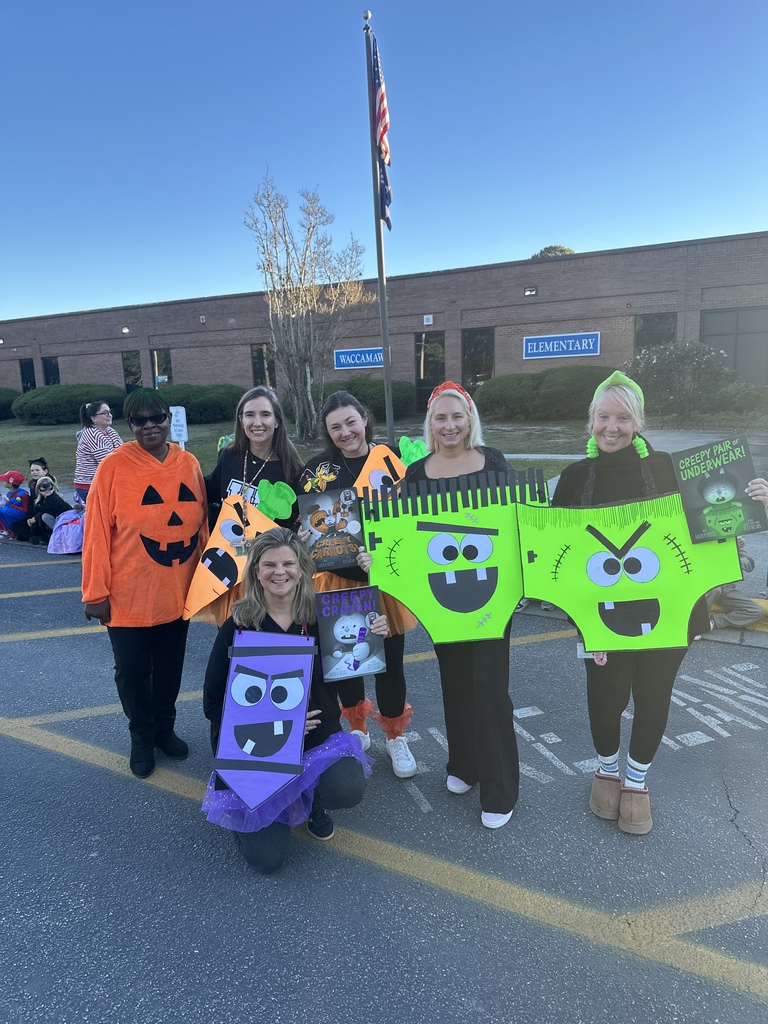 Six teachers stand outside Waccamaw Elementary in front of the American flag and school sign, dressed in Halloween-themed costumes inspired by Creepy Carrots and Creepy Pair of Underwear. Some wear pumpkin shirts, others hold large colorful cutouts shaped like underwear and crayons, and one teacher kneels in front holding a Creepy Crayon book.